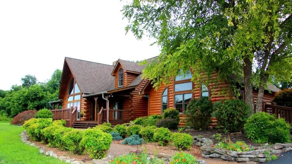 Photo of log home with round log exterior, upper dormers and vaulted great room with geometric windows floor to ceiling. A vaulted paledium window creates height to the home's right side wing