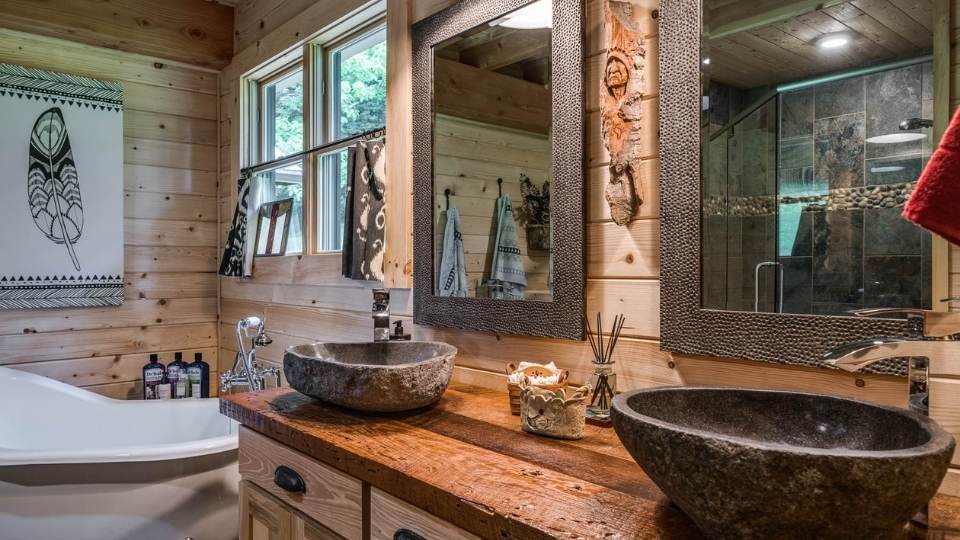 Photo of a log home bath with double above counter sinks and a soaker tub. The walls are exposed light color wood that contrasts with the dark wood countertop of the vanity