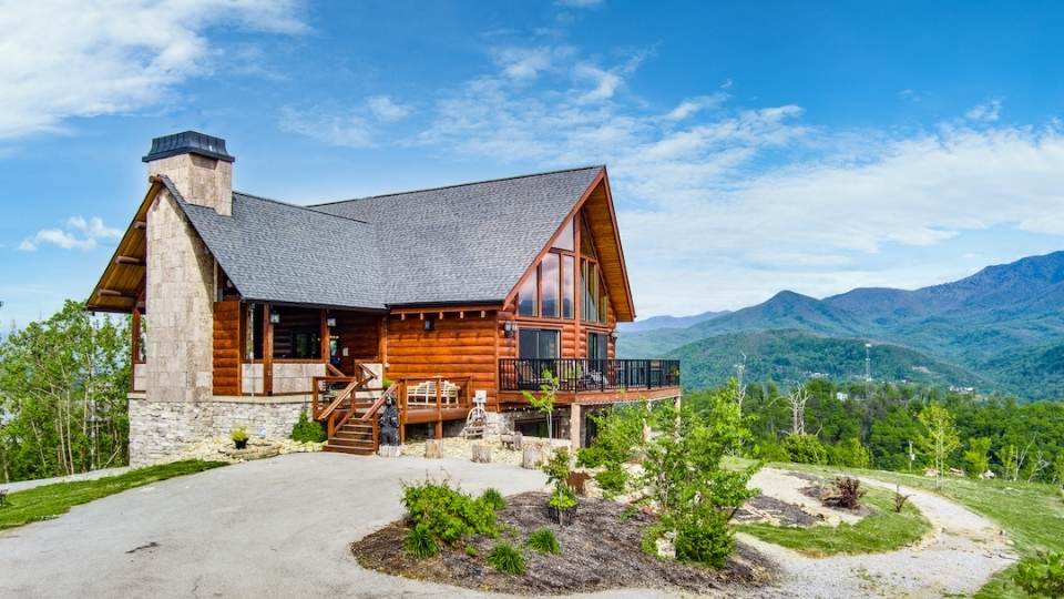 Photo of log home with a massive stone fireplace on the screened porch. The photo is an exterior shot showing the stunning view of a valley and mountains. The home's great room sports two stories of geometric glass and glass doors leading to an exterior deck