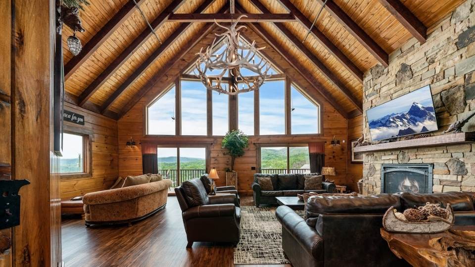 Photo of a great room with the interior wall of stone with a fireplace in the center at ground level. The vaulted ceiling is exposed wood with exposed wood dark brown beams. The exterior wall is floor to ceiling glass with the ground level providing sets of glass doors leading to an exterior deck.