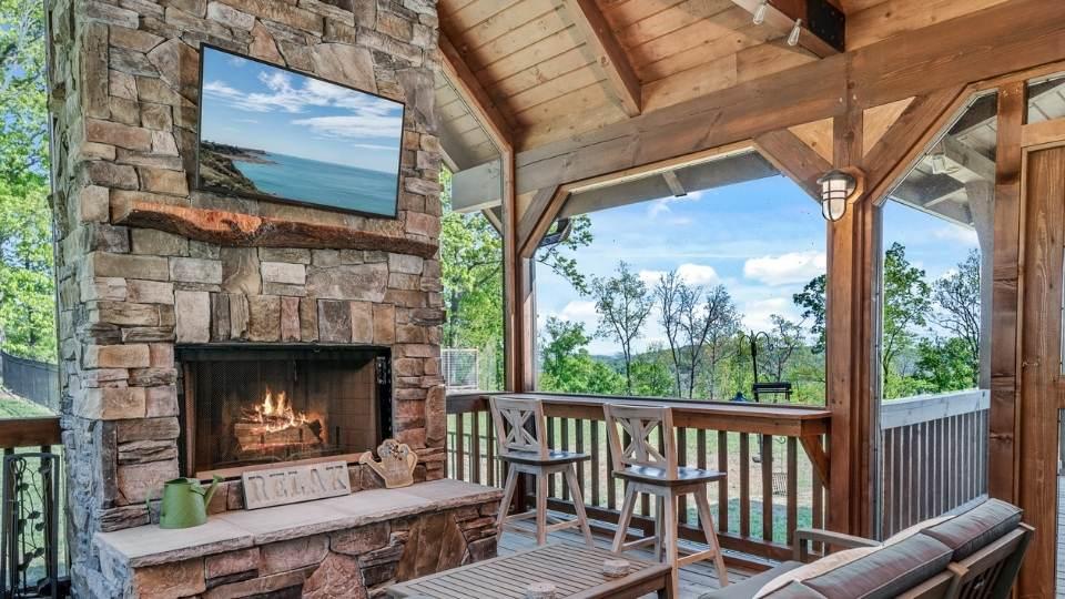 Photo of outdoor living porch with built in bar seating on the porch raliing. A two story stone fireplace with a tv over the mantel creates a sense of a family room