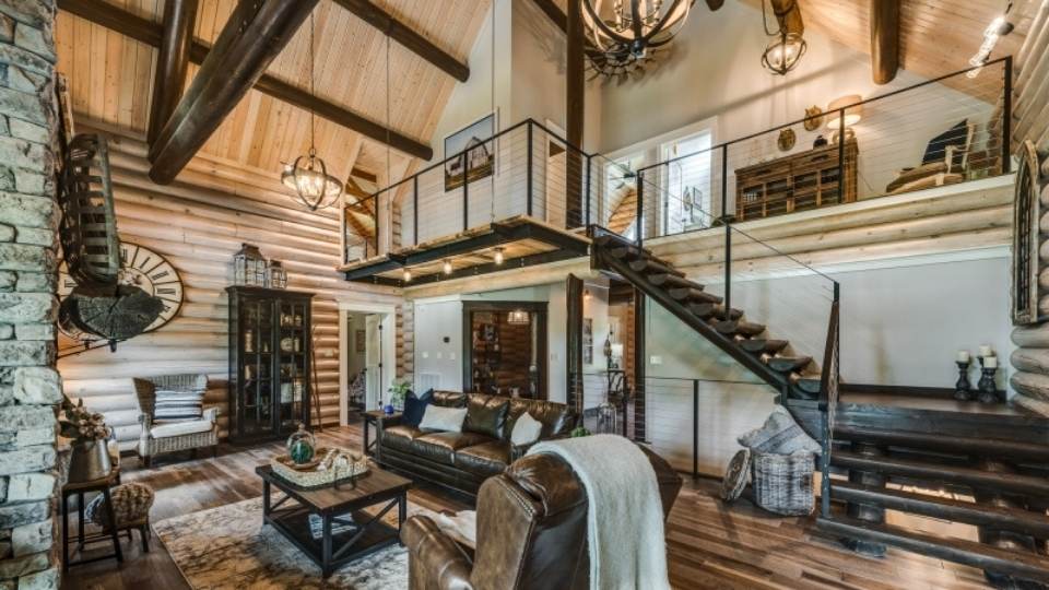 Photo of great room of timber frame home with exposed dark wood stairs leading the the open loft/balcony. the stair case uses wire rails creating a competely open view. the great room walls are exposed round wood and the ceiling is exposed honey oak with contrasting dark wood supports